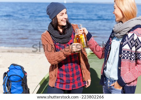 Cheers to us! Two beautiful young women cheering with beer and smiling while standing near their tent on the beach