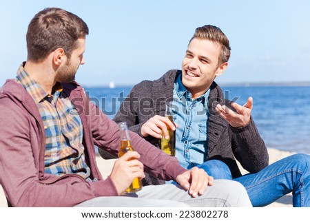 Taking time to talk with friend. Two handsome young men drinking beer and talking to each other while sitting on the beach together