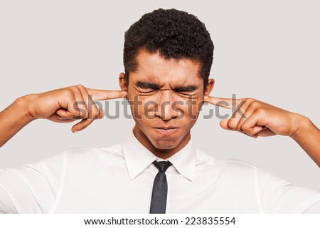This is too loud! Frustrated young Afro-American man in formalwear plugging ears with his fingers and keeping eyes closed while standing against grey background