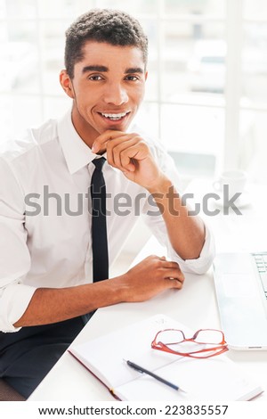 Pleasant working! Handsome young Afro-American man in shirt and tie holding hands on chin and smiling while sitting at his working place