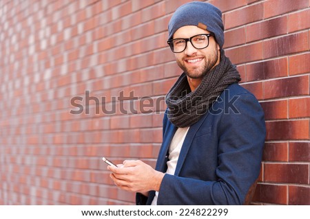 Typing text message. Side view of handsome young man in smart casual wear holding mobile phone while leaning at the brick wall