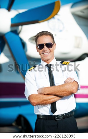 My job is my passion. Confident male pilot in uniform keeping arms crossed and smiling while standing in front of the airplane