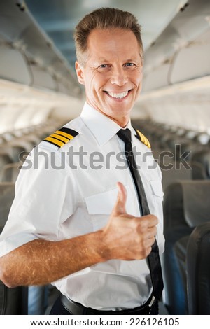 I love my job! Confident male pilot in uniform showing his thumb up and smiling while standing inside of the airplane