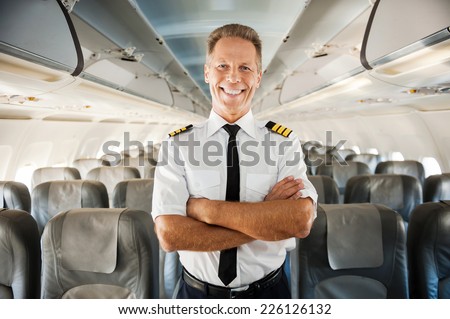 This is my plane. Confident male pilot in uniform keeping arms crossed and smiling while standing inside of the airplane