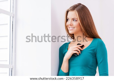 Waiting for your call. Beautiful young woman holding phone in her hand while standing near window