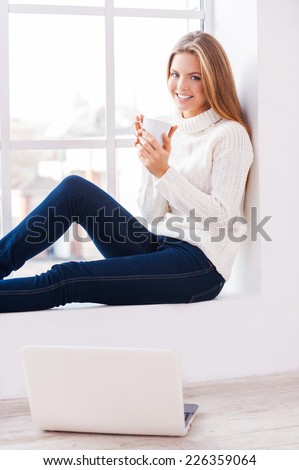 At home warm and comfortable. Beautiful young woman sitting near the window at her apartment holding a cup of coffee