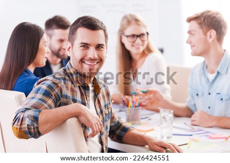 Happy to be a part of creative team. Group of cheerful business people in smart casual wear sitting together at the table and discussing something while handsome man looking at camera and smiling