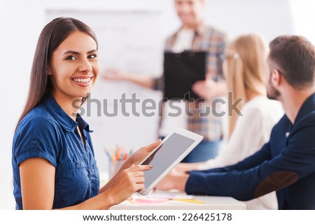 Happy to be a part of team. Confident young woman holding digital tablet and smiling while man standing in the background and pointing whiteboard