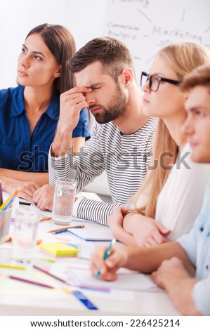 Tired and stressed. Depressed young man touching face with hand while sitting at the table together with his colleagues