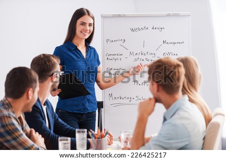 Feeling confident in her speech. Group of business people in smart casual wear sitting together at the table while beautiful woman standing near whiteboard and pointing it with smile