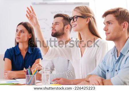 I have a question! Group of business people in smart casual wear sitting together at the table and looking away while beautiful woman raising her hand up and smiling