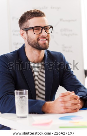 Confident business expert. Confident young man in smart casual wear looking away and smiling while sitting at his working place with whiteboard in the background
