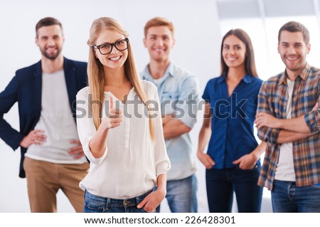 Join a successful team! Beautiful young woman showing her thumb up and smiling while group of happy young people standing on background and smiling