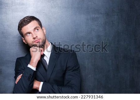 Thoughtful businessman. Handsome young man in shirt and tie looking away and holding hand on chin while standing against blackboard