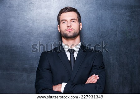 Confident business. Handsome young man in shirt and tie looking at camera and keeping arms crossed while standing against blackboard