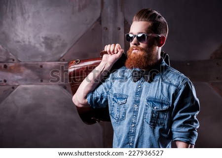Stylishly confident. Handsome young bearded man in sunglasses looking away while holding a suitcase on his shoulder