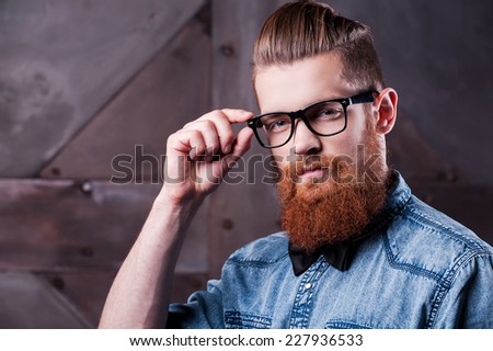 Being retro is a trend. Handsome young bearded man holding hand on glasses and looking at camera