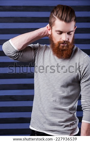 Casual and cool. Side view of handsome young bearded man holding hand on head while standing against stripped background