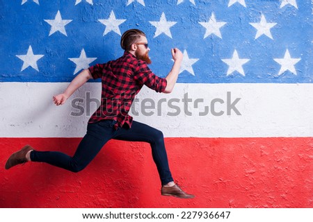 Hurrying to be in trend. Handsome young bearded man wearing sunglasses and running along American flag