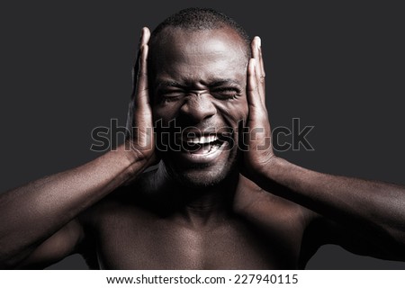 Too loud sound. Portrait of young shirtless African man covering ears with hands and shouting while standing against grey background