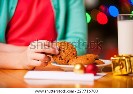 My favorite Christmas cookies! Close-up of little girl taking a cookie from plate while sitting at the table with Christmas Tree in the background