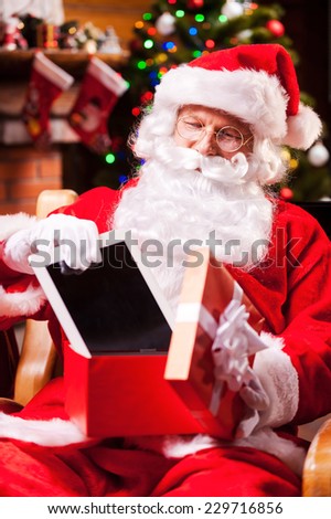 Good present! Cheerful Santa Claus putting a digital tablet into the gift box and smiling while sitting at his chair with Christmas Tree in the background