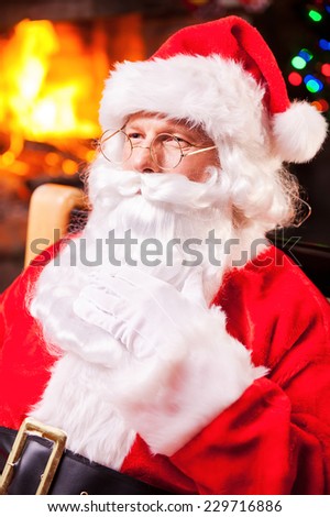 Thoughtful Santa. Thoughtful Santa Claus sitting at his chair and touching beard with fireplace and Christmas Tree in the background