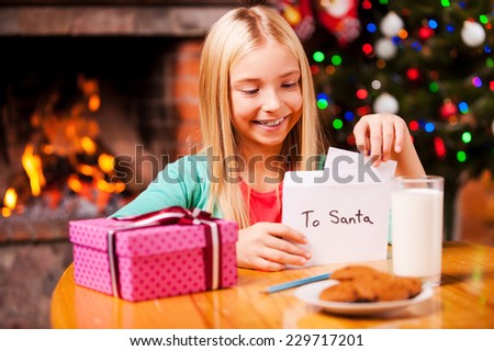 Wish list to Santa. Cute little girl putting a letter to Santa into the envelope while sitting at home with Christmas tree and fireplace in the background