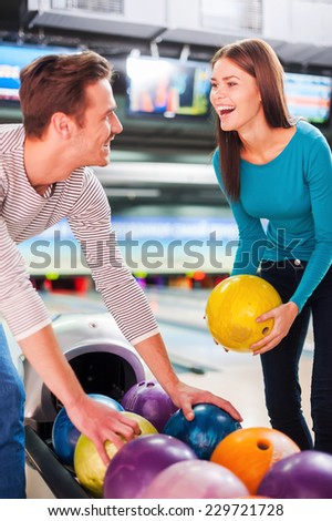 Cheerful friends. Cheerful young couple looking at each other and choosing bowling balls while standing against bowling alleys