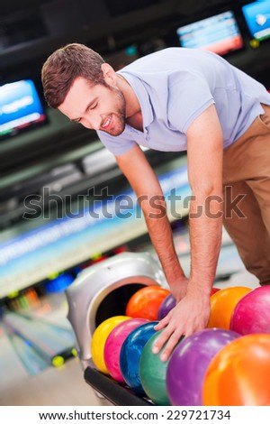 Choosing the lucky ball. Cheerful young man choosing bowling ball and smiling while standing against bowling alleys