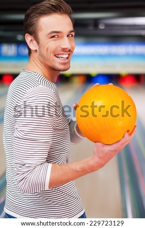 Hoping for a strike. Cheerful young man looking over shoulders and holding a bowling ball while standing against bowling alleys