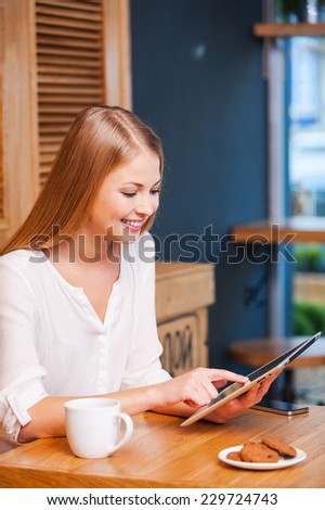 Enjoying a digital age. Side view of beautiful young woman using digital tablet and smiling while enjoying coffee in cafe