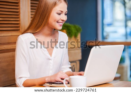 Working with pleasure. Side view of beautiful young woman working on laptop and smiling while enjoying coffee in cafe