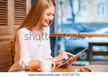 Surfing the net in cafe. Side view of beautiful young woman using digital tablet and smiling while enjoying coffee in cafe