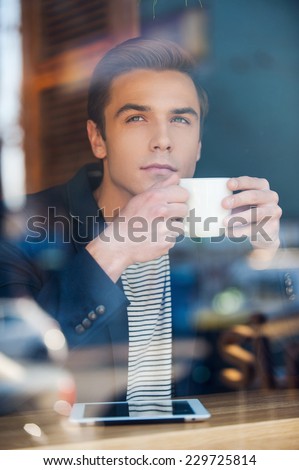 Waiting for inspiration. Through a glass shot of handsome young man writing something in his note pad while enjoying coffee in cafe