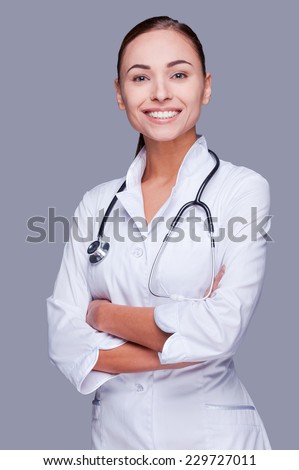 Taking good care of your health. Confident female doctor in white uniform looking at camera and smiling while standing against grey background