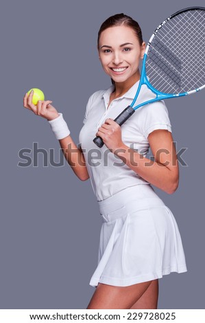Bringing the beauty to the game.  Beautiful young women in sports clothes holding tennis racket on her shoulder and smiling while standing against grey background