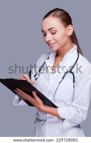 Beautiful doctor at work. Confident female doctor in white uniform writing clipboard while standing against grey background