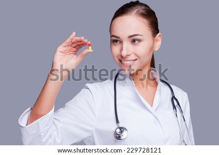 Take this pill! Female doctor in white uniform holding pill and smiling while standing against grey background