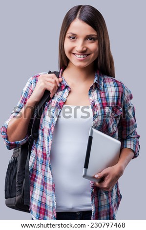 New age for studying. Happy young women carrying a backpack on her shoulder and holding digital tablet while standing against grey background