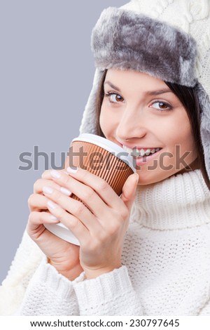 It is time for warm drinks . Close-up of young women in warm winter clothing drinking coffee and smiling while standing against grey background