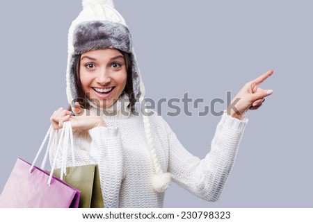 Incredible time for purchases. Happy young women in warm winter clothing holding packages with purchases and pointing away while standing against grey background
