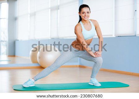 Woman exercising. Beautiful young woman in sports clothing doing stretching exercise and smiling while standing in health club