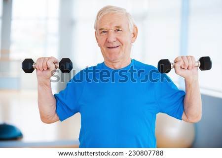 Staying healthy. Happy senior man exercising with dumbbells and smiling while standing indoors