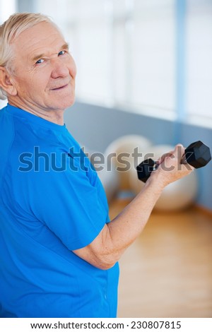 Staying active and healthy. Rear view of happy senior man exercising with dumbbells and looking over shoulder while standing in health club