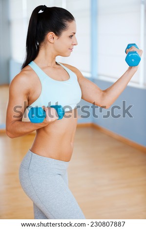 Exercising with dumbbells. Side view of beautiful young woman in sports clothing exercising with dumbbells and smiling while standing in health club