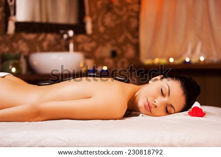 Total relaxation. Side view of beautiful young shirtless woman lying on massage table and keeping eyes closed with spa stones on her back