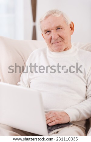 Surfing the net. Confident senior man working on laptop and looking at camera while sitting in chair at his apartment
