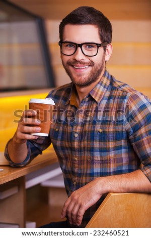 Enjoying hot and fresh coffee. Happy young man holding coffee cup and smiling while sitting in coffee shop