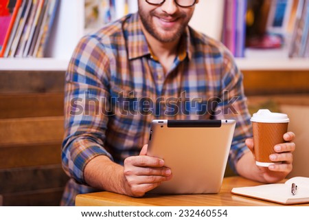 Digital age student. Close-up of cheerful young man holding coffee cup and looking at his digital tablet while sitting at the desk with bookshelf in the background